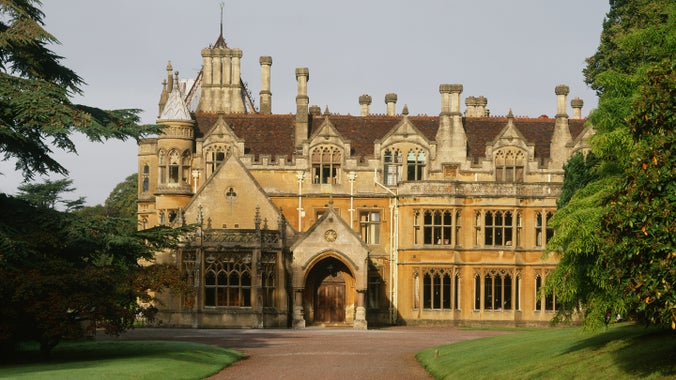 A view along a driveway to the front of a large country house, in a gothic style with tall chimneys and triangular gables along the roof